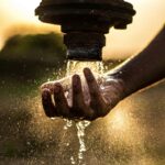 A person washes their hand under a rustic water pipe, capturing the essence of fresh water at sunset.